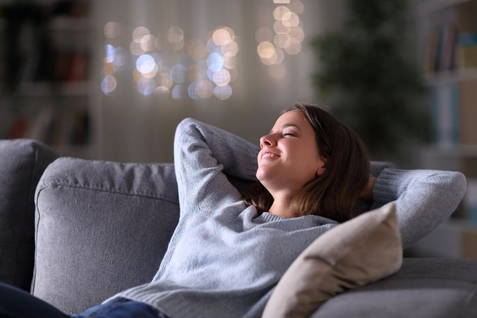 A white woman with straight brown hair reclines on a coach with her eyes closed. She is smiling. In the blurred-out background, there is a Christmas tree.