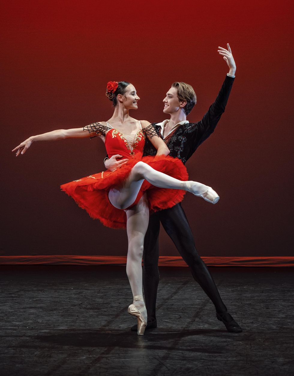 A white ballerina in a bright red tutu, pink tihgst and pointe shoes and a rose in her hair performs an attitude devant with her right leg and looks towards her partner on her left. Her partner a white male danseur, wears a black bolero jacket, black tights and slippers and holds her around the waist with his right hand with his left arms up high.