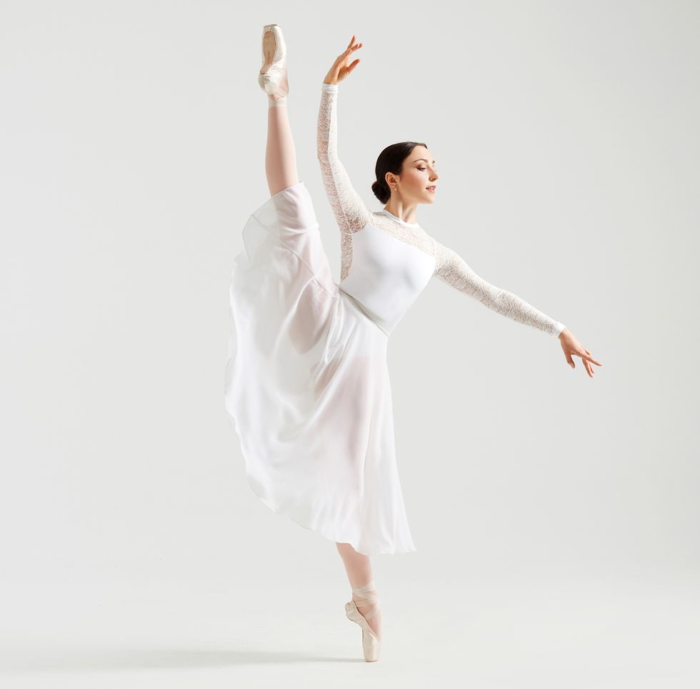 Paraschiv is dressed in a white lace long-sleeved leotard and long white leotard. She's in a high du00e9veloppu00e9 to the site with her gaze over her lower hand.
