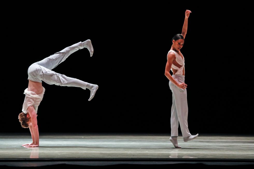 A male dancer in a white shirt and pants begins to do a handstand while a female dancer to his right steps high on demi pointe on her left foot and lifts her left arm above her head.
