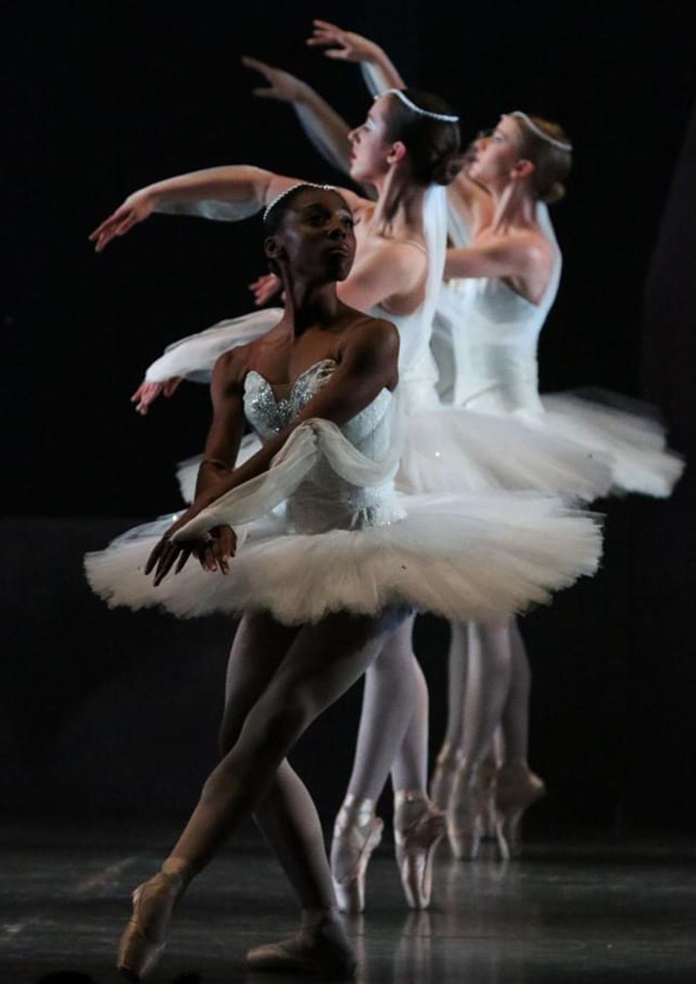 Aerys Merrill is at the front of a line of dancers in classical white tutus and pointe shoes. She is in pliu00e9 tendu devant while the dancers behind her stand in sous-sus.