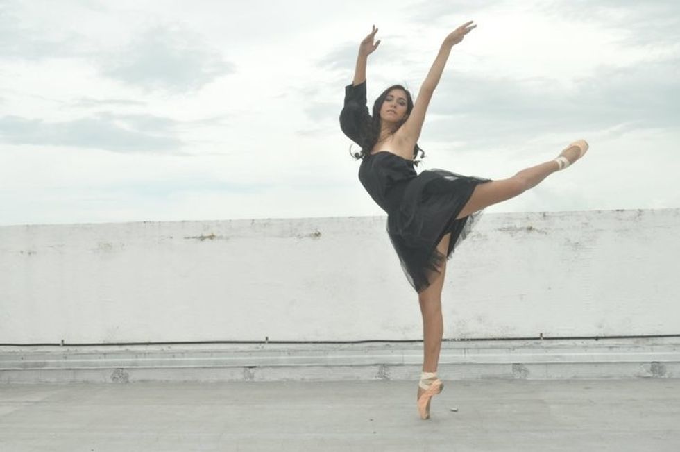 A young woman with long, dark hair and wearing a black dress, poses on a rooftop in piquu00e9 attitude, her arms high in a v-shape.