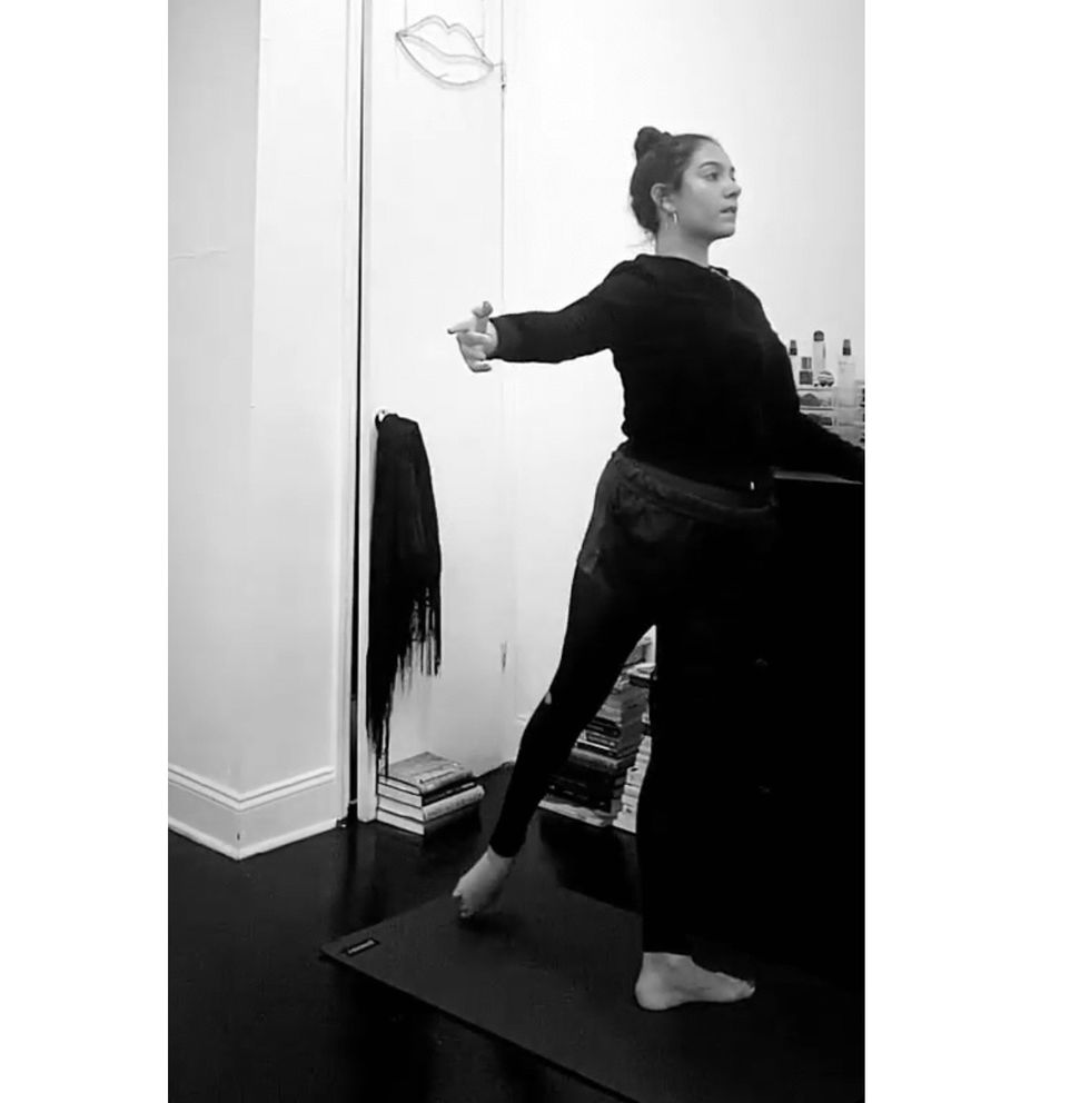 A young woman with her hair in a bun and wearing dark dance clothes holds onto a bedroom dresser and performs tendu derriu00e9re.