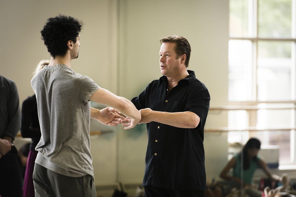 Wearing a short-sleeved black button-down shirt, Mikko Nissinen presses his hands against a male dancer's arms held in first position during a company class at Boston Ballet.