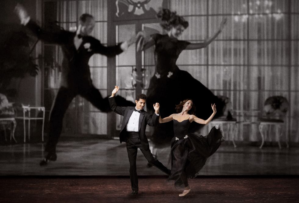 Renan Cerdeiro and Ashley Knox, holding hands, leap lightly onto their right feet, their arms thrown up and their left leg kicked up beside them. He wears a black tuxedo and she wears a strapless black gown. Behind them onstage looms a giant black-and-white projection of Fred Astaire and Rita Hayworth, wearing similar costumes.