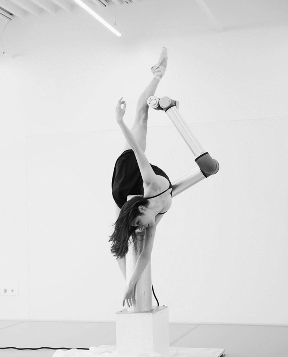 Merritt Moore drapes her upper body over the base of a robot and extends her right leg high in the air. The robot arm rests against her lifted leg.
