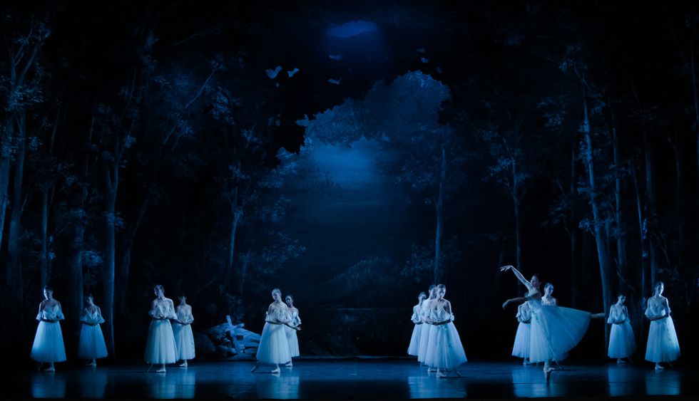 On a darkened stage with a forest backdrop, a group of 13 ballerinas in long white tutus stand in B+ with their lower arms crossed at their waist. Downstage right, a soloist dancer does a piquu00e9 third arabesque on her right leg.