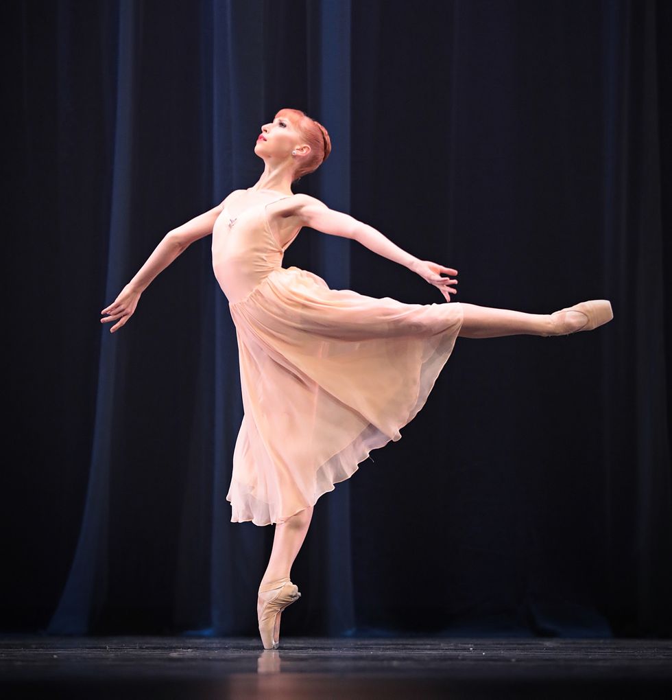A dancer in a pink chiffon dress with red hair in a bun stands in a 90-degree arabesque, with her arms stretched out.
