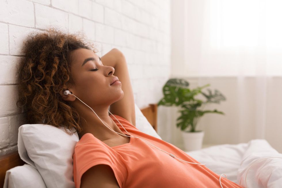 A young Black woman in a coral-colored T-shirt lays back on a pillow with her eyes closed. She looks peaceful and wears ear buds as sun filters into the room through filmy curtains.