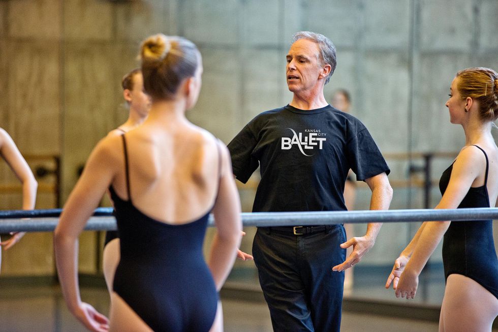 Devon Carney, an middle-aged ballet master with gray hair and a black T-shirt and jeans, demonstrates fifth position with his arms en bas. In front of him are a few teenage ballet students in black camisole leotards, mimicking his position.