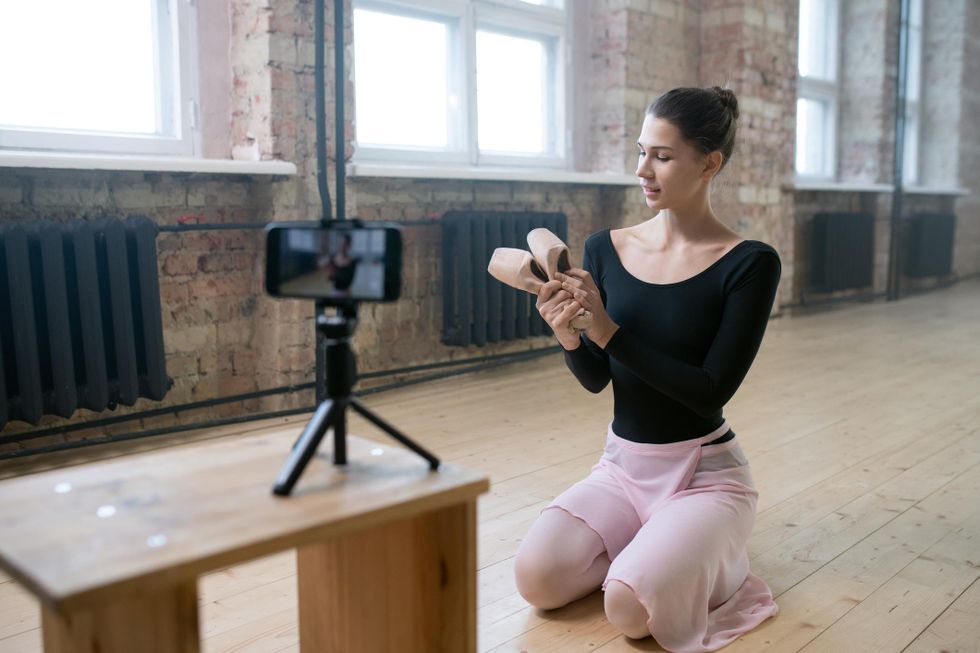 A young ballerina in a black leotard and pink skirt kneels on the wood studio floor and holds up a pair of pointe shoes. In front of her, a smart phone camera on a tripod is propped on a wooden stool.