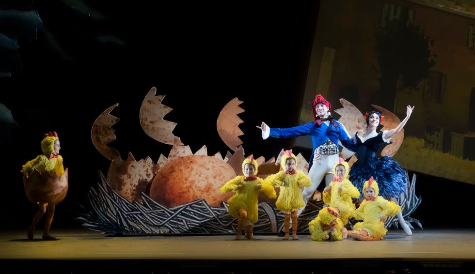 Jacob Bush, in a blue jacket and red hat, stands next to Jackie Nash, who wears a black and blue tutu, both in tendu back. To their left is a nest full of large broken eggs, along with six child dancers dressed as yellow chicks.