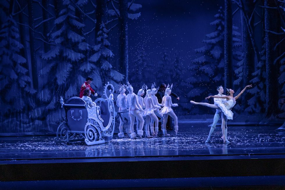 A ballerina in a white tutu poses in arabesque, partnered by a male dancer dressed in a white jacket and crown. To their left, a group of six dancers dressed as white reindeer pull a sleigh towards a wintery backdrop; a male dancer dressed as a Nutcracker prince stands on the sleigh and a young female dancer sits inside it.