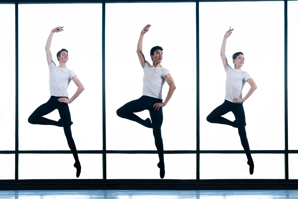Three teen boys in black tights and white shirts jump in front of large windows in a studio. They have one leg in passe, one arm in fifth position and the other hand on their hips