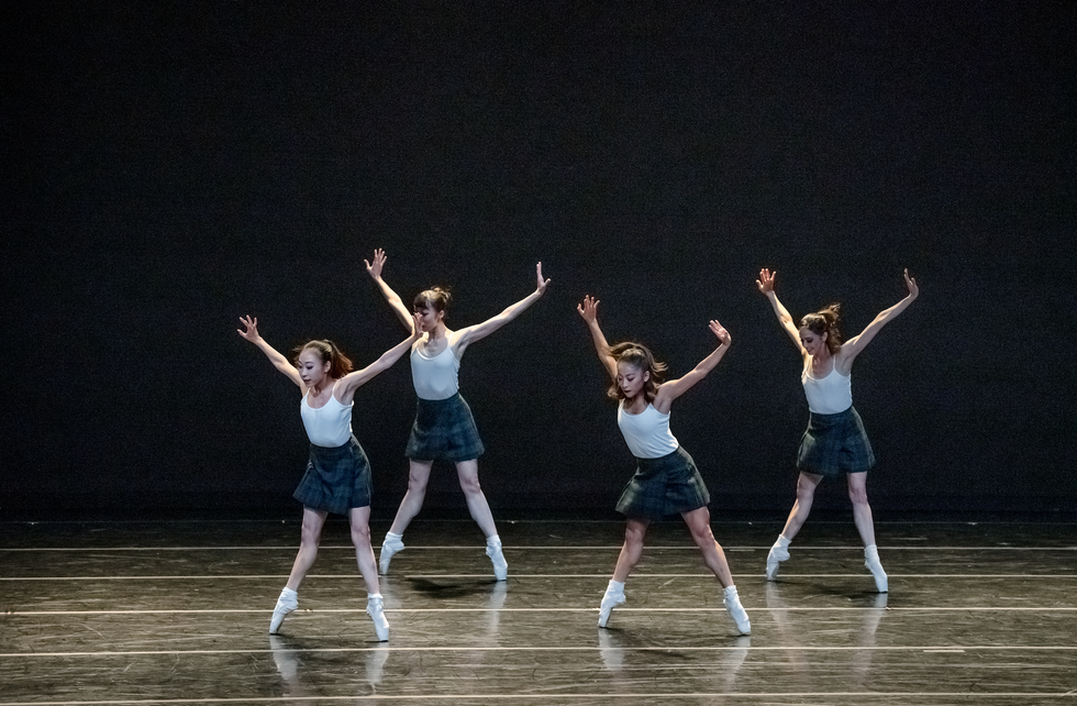 Four dancers onstage in white leotards, plaid skirts and white socks and pointe shoes, standing in second position leaning over.