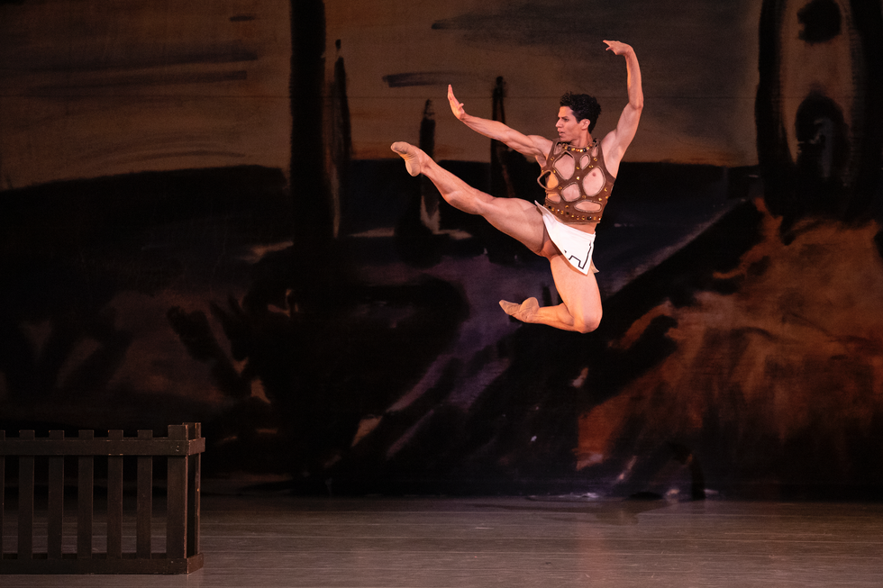 Diniz leaps in the air against a brown painted backdrop onstage, in a gold and brown mesh top and white grecian skirt.