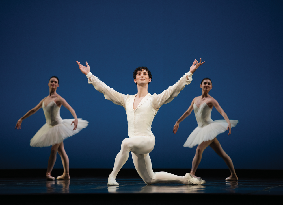 Cauthorn in all white kneels onstage with his arms int he air. Ballerinas dressed in white stand in a fourth position on either side of him.