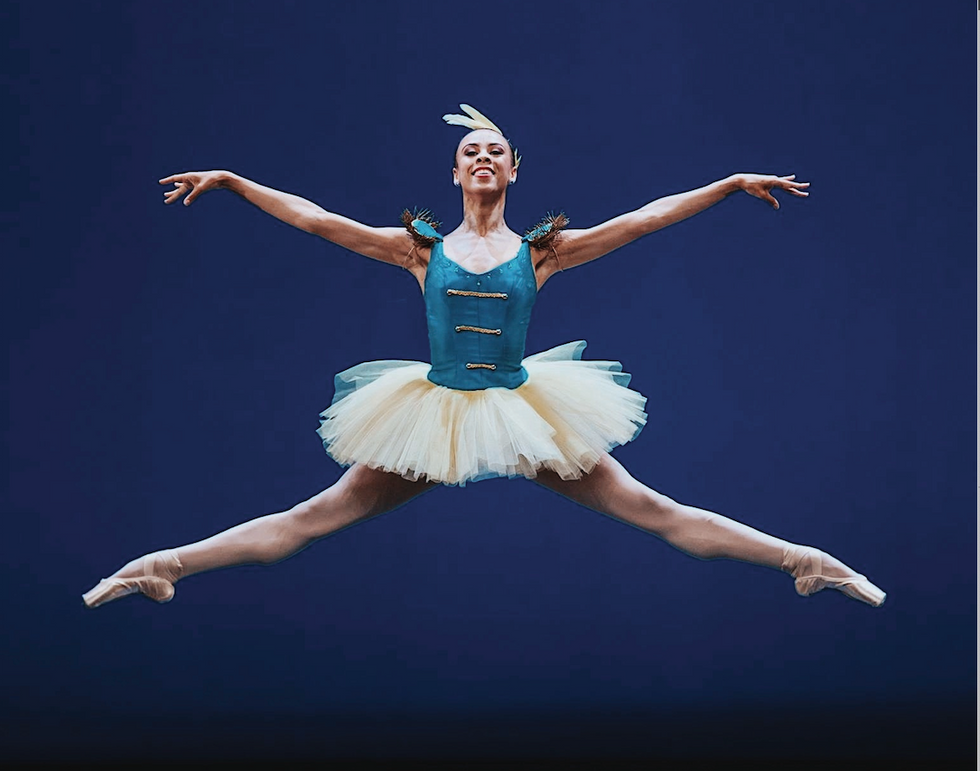 Fogo, in a blue military bodice and yellow tutu, jumps in the air with both toes pointed.