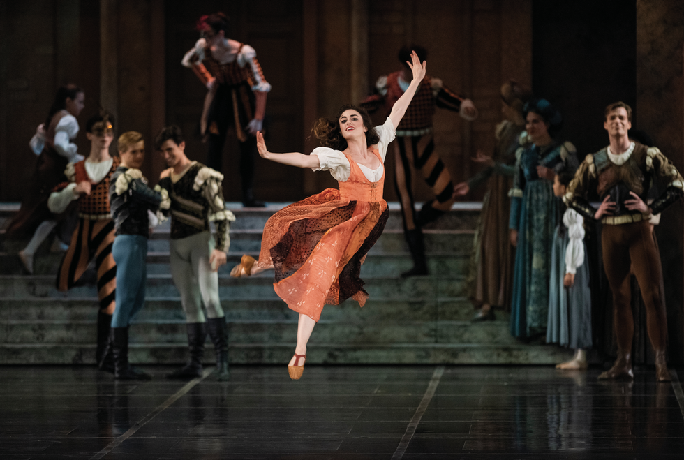 Ellen Rose Hummel in an orange peasant dress, white blouse and heeled shoes, jumps in the air with her hands flexed and dark hair loose.