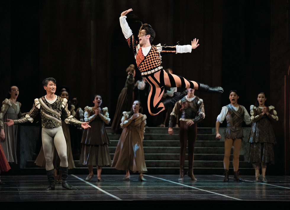 Erni leaps high in the air with one leg in passu00e9 in an orange and black jester like outfit, while dancers dressed as peasants around him look on in shock.