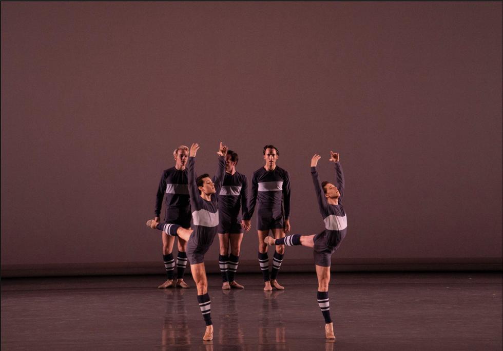 Two male dancers in blue shorts, a blue long-sleeved shirt with a large gray stripe, and blue and gray striped legwarmers, do piquu00e9 arabesque with their arms in high fifth. Three men in similar costumes stand casually behind them onstage.