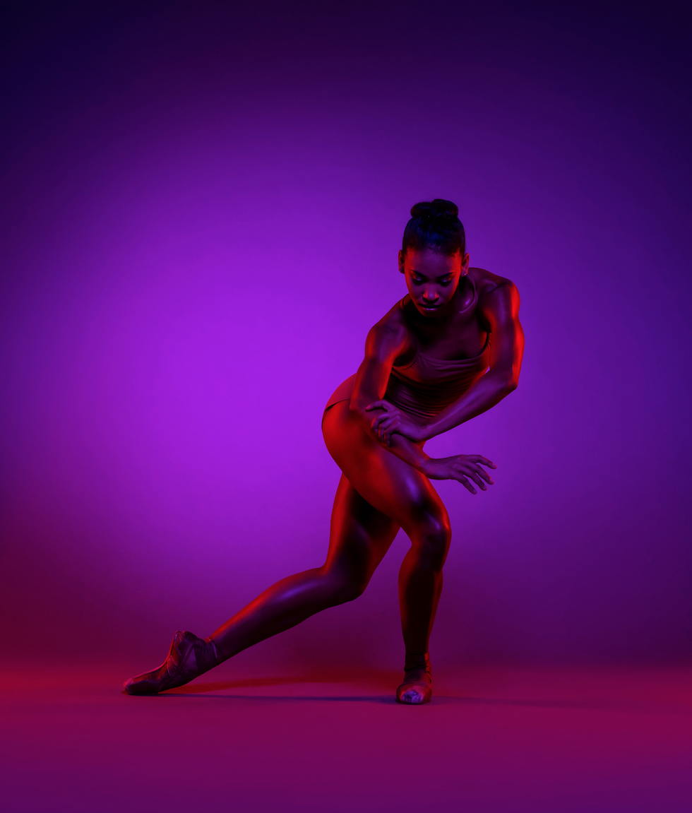 In a moody purple light, Raven Barkley twists and steps to the left on a bent right leg, crossing her left leg behind her and grabbing her right forearm with her left hand.