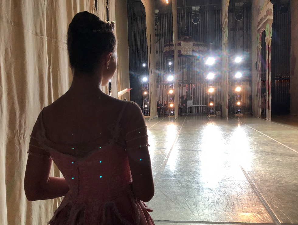 The silhouette of ballerina standing backstage in the wings is shown from the waist up; she wears a long pink tutu and faces bright boom lights in the wings across the stage.