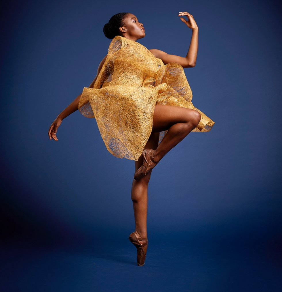 In front of a dark blue background, Wimpye, wearing abstract corky material, is en pointe with her right leg in passu00e9.