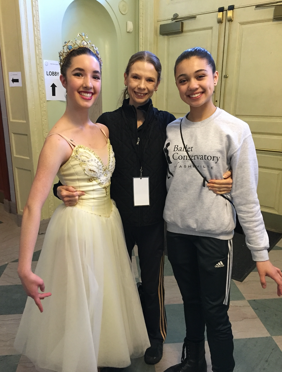 A female ballet teacher in black athleisure wear poses in the middle of two female ballet students, holding their waists and smiling for the camera. The dancer on her left wears a long white tutu and crown, while the dancer on her right wears black workout pants and a gray sweatshirt.