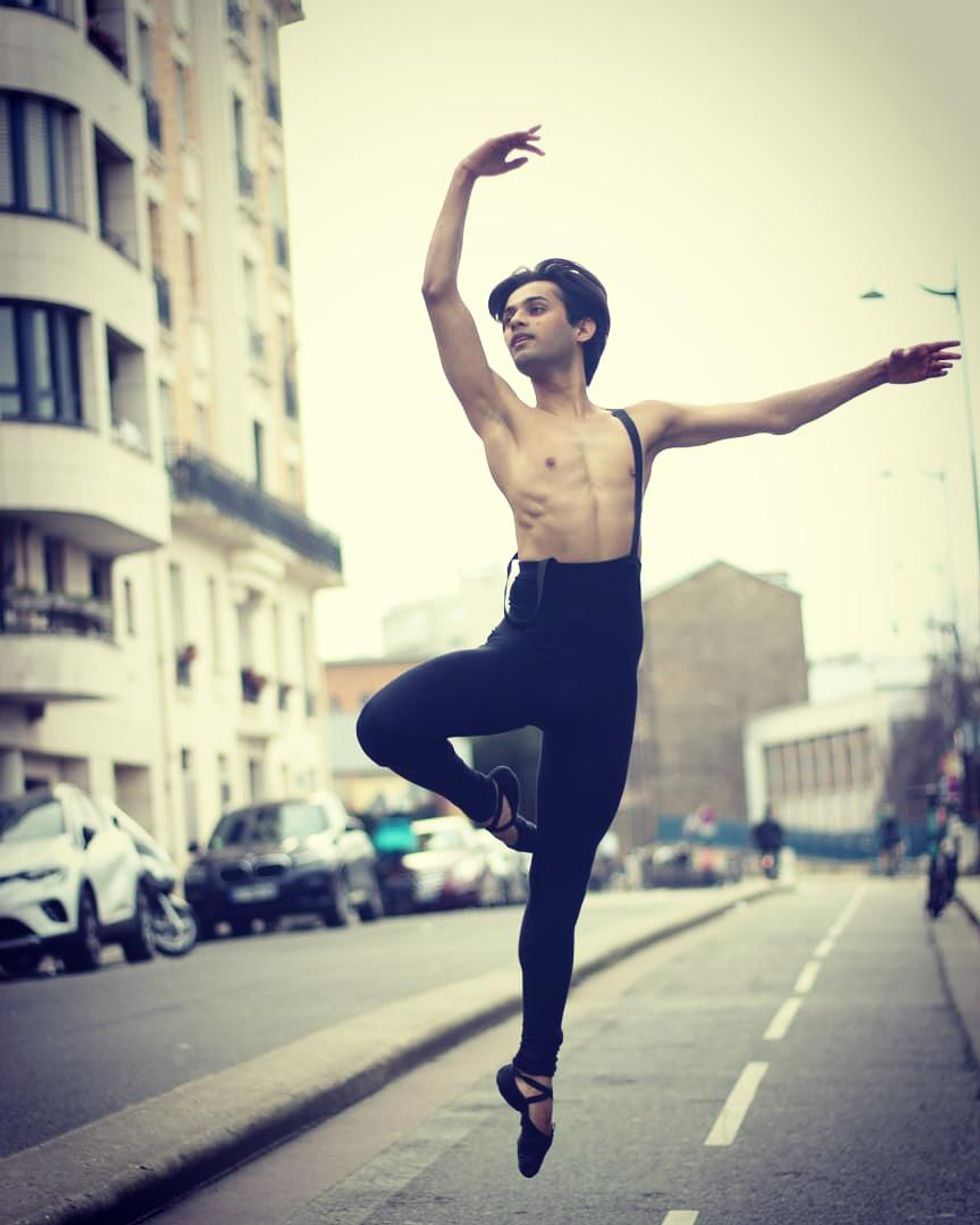 Bobby Roy is photographed midair, jumping in passu00e9 in a city street scene. He is wearing black ballet shoes, and black tights with on suspender over his bare chest.