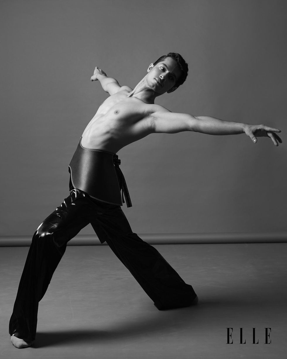 A black and white image of Manish Chauhan in a fourth position lunge with arms in second as he arches his torso backwards. He is photographed bare-chested and in shiny, high-fashion pants.