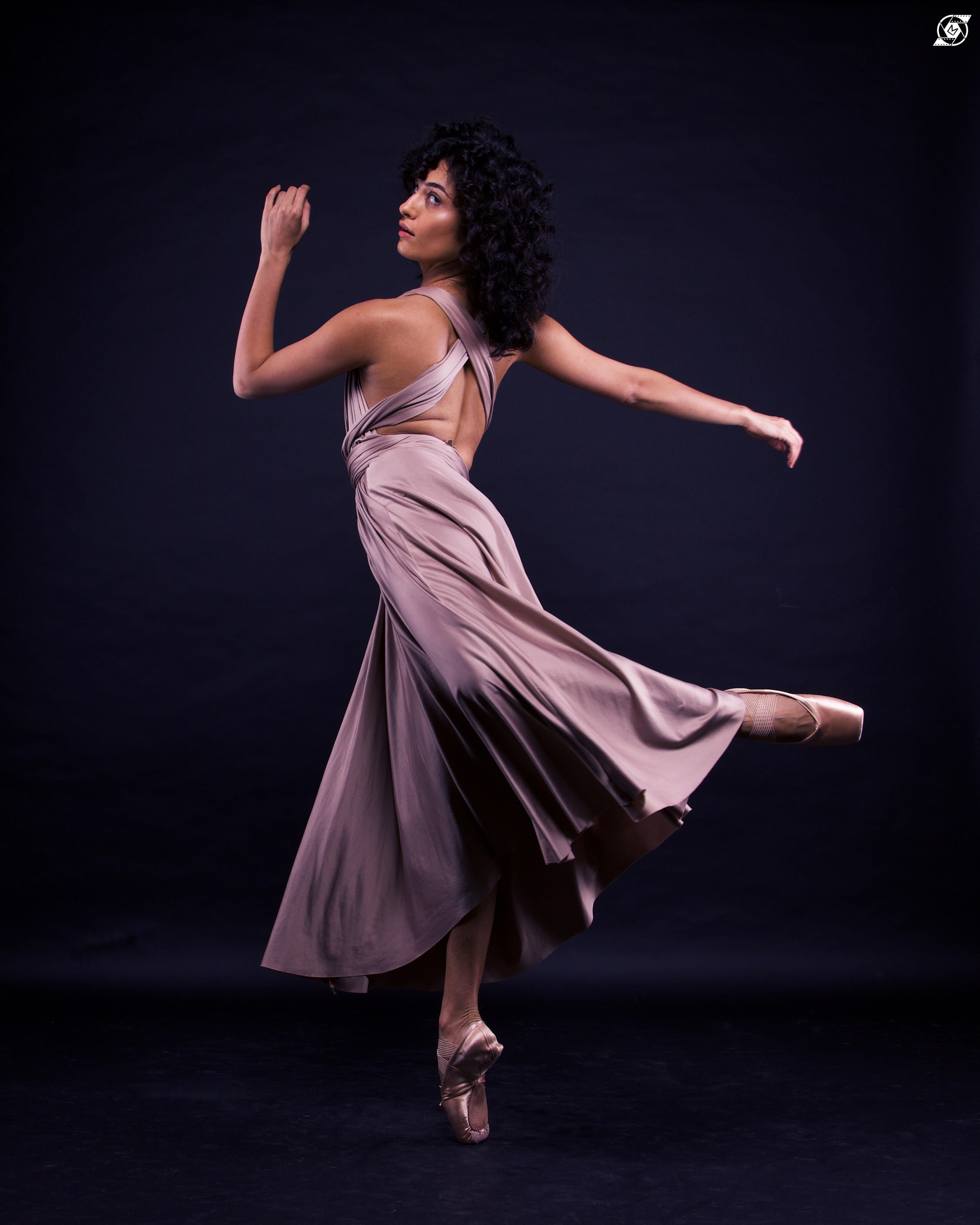 Pia Sutaria, in pointe shoes and a flowing lavender dress, does a piquu00e9 in low attitude derriu00e8re as she glances over her left shoulder at the camera. Her dark curly hair falls over her shoulders.