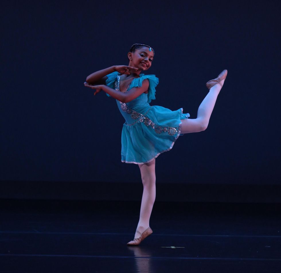Vidhi Thakker, an 11-year-old Indian girl in a bright blue ballet costume and tights, performs onstage. She poses in piquu00e9 attitude with her arms framing her face.