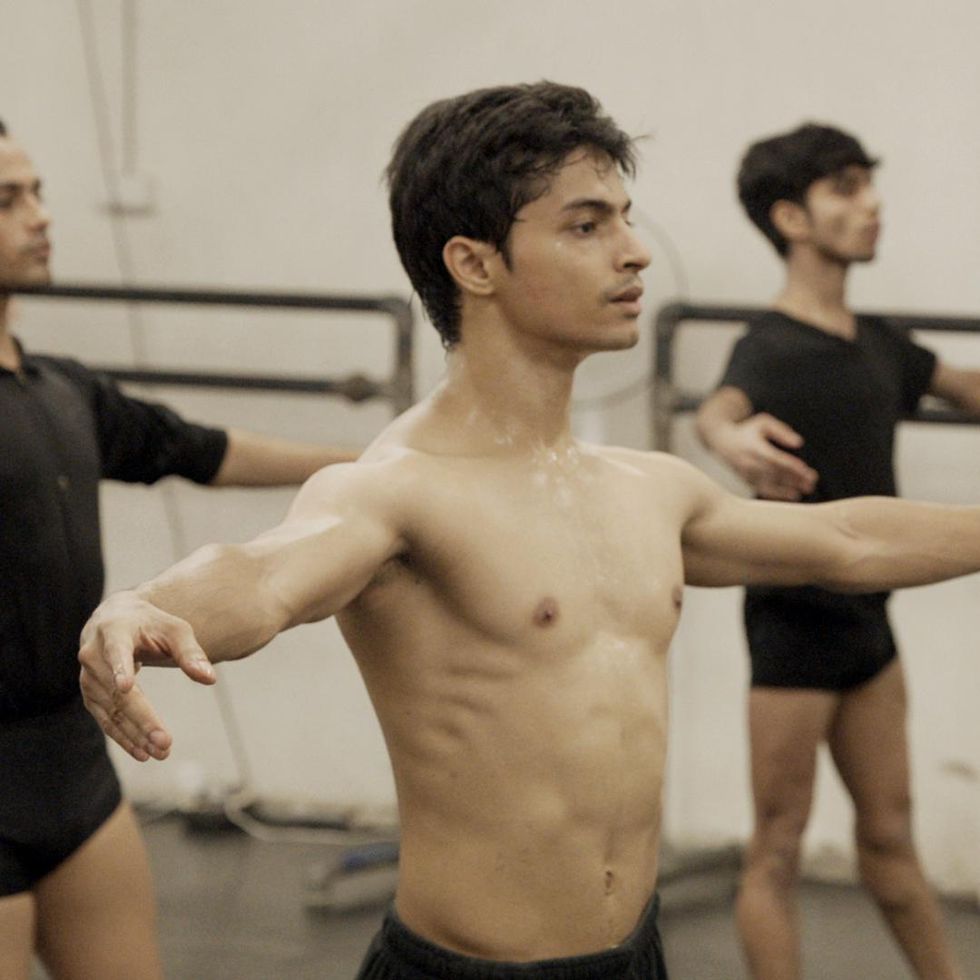 Three young Indian men do tendus in a ballet class. One is sweating with no shirt on. The other two men, behind him are wearing black shirts and shorts.
