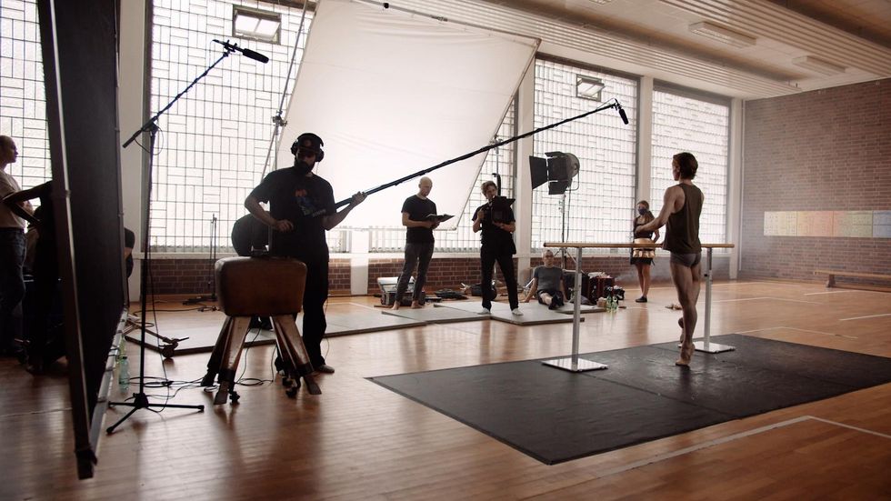In a gymnasium set up with large boom lights and video cameras, Daniil Simkin balances in coupu00e9 a the barre with his back away from the camera. He wears gray booty shorts, a green tank top and tan ballet slippers. Three crewmen and a woman in a patterned skirt stand in front of him.