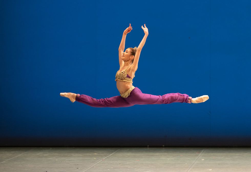 Weearign a gold bra top, purple harem pants and pointe shoes, and young female ballerina does grand jeté through the air onstage with her arms in high fifth position.