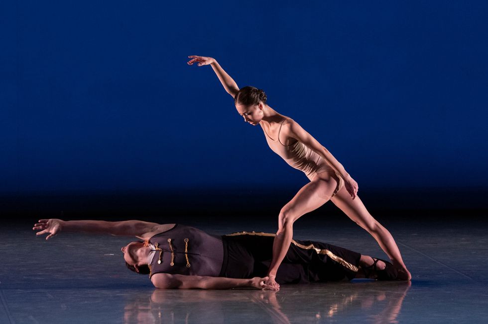 A male dancer in a gray costume lies on the floor and reaches his left hand out above his head. A female dancer in a tan leotard stands over him in a wide fourth position.