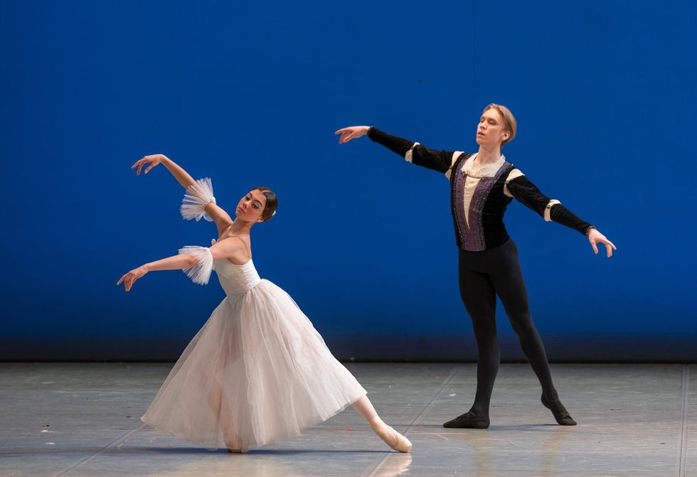 A ballerina in a white Romantic tutu pliés in tendu derriere effacé, her arms in third arabesque position. Behind her, a young male danseur in a black tunic and tights, does tendu derriere effacé on hus right leg and holds his arms out to the side.