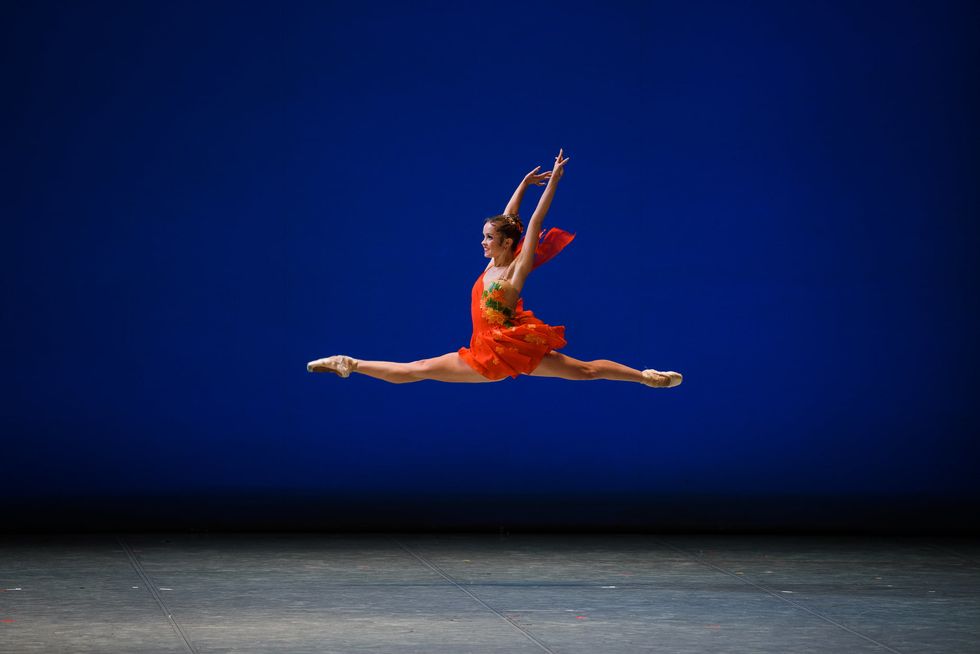 A young ballerina in a red tunic costume, pink tights and pointe shoes leaps through the air onstage with her arms thrown high above her head.