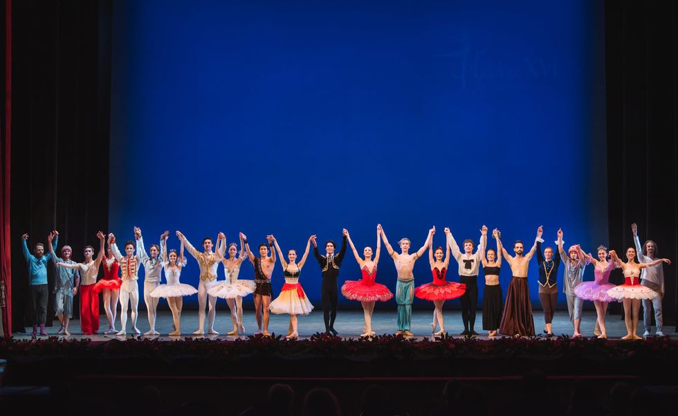 A long line of male and female ballet dancers in various costumes stand at the edge of the stage, hold hands and raise their arms up above them in preparation for a bow.