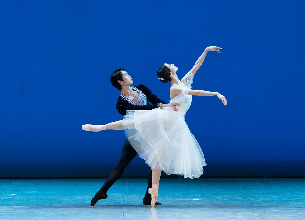 A male danseur wearing a black velvet jacket and black tights holds a ballerina's waist and leans back behind her, his right leg in tendu. The ballerina wears a white romantic tutu and stands on her left leg in a low third arabesque, looking up towards her left hand.