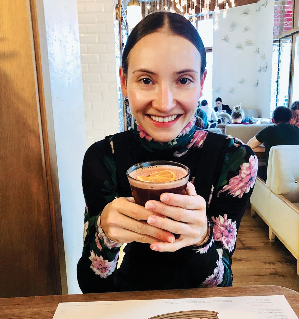 Shevchenko smiles in a cafu00e9 while holding a hot beverage, wearing a flowered turtleneck.