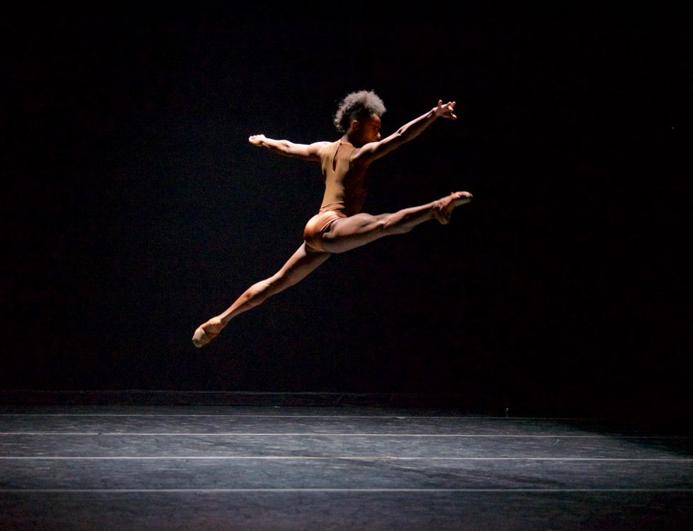 A black dancer flies through the air in a split on a black stage
