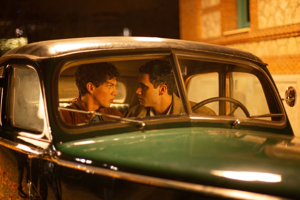 Two men in brown jackets are shown talking closely in the front seat of a green 1950s-style car.