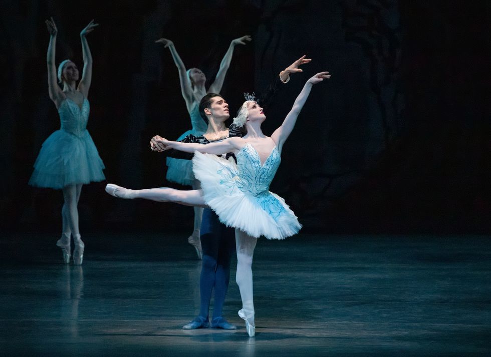 Ashley Bouder, in a white swan tutu, poses in first arabesque on pointe while Jovani Furlan, in dark tights and a black tunic, stands behind her and holds her right hand.