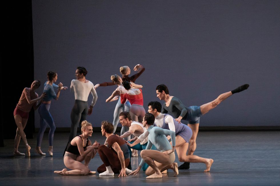 A group of dancers in multi-colored costumes form a clump. Some crouch or kneel on the ground, while Jovani Furlan stakes arabesque.