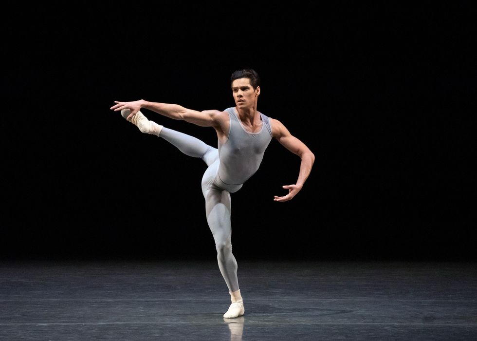 Jovani Furlan, wearing a gray unitard and off-white socks and slippers, stands in attitude croisu00e9 with his right (standing) leg in pliu00e9. His right arm is stretched out to the side and his left arm is curved.