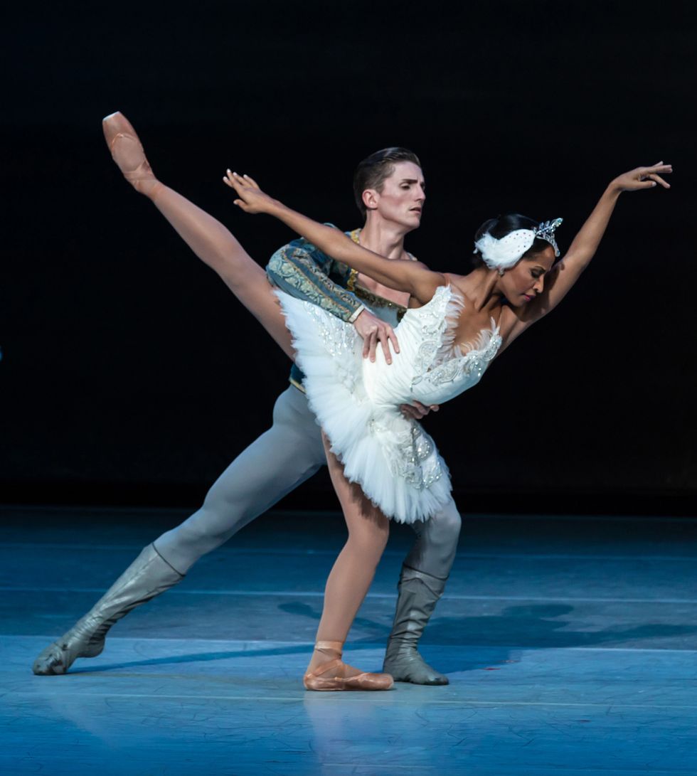 Kayla Rowser, costumed in a white swan tutu, pliu00e9s in first arabesque allongu00e9 on her left leg. Nicholas Scheurer, in a blue-green price costume, holds onto her waist behind her.