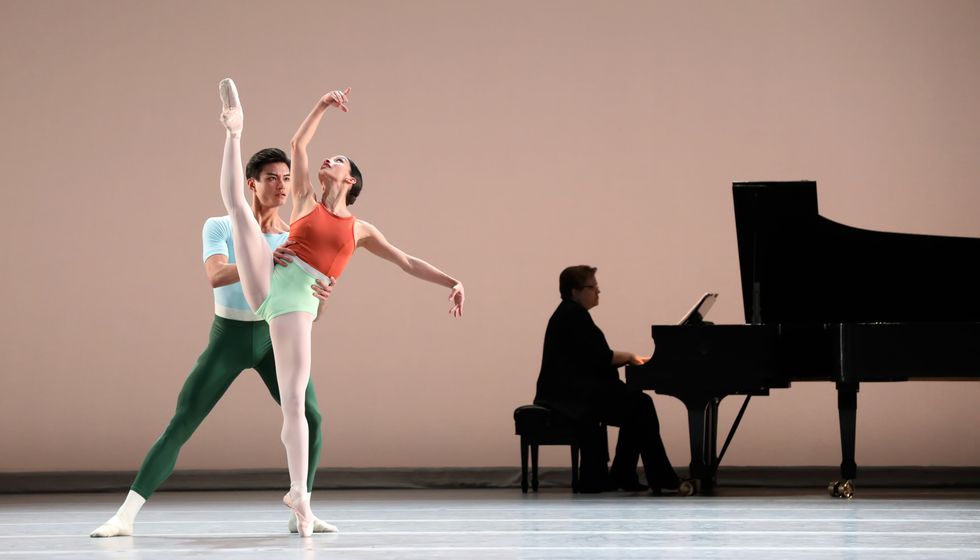 Wearing an orange and green leotard, pink tights and pointe shoes, Karina Gonzu00e1lez extends her right leg high to the side and looks up towards her right hand, which is lifted. Chun Wai Chan, in dark green tights and a mint T-shirt, holds onto her waist while a male piano player sits at a piano upstage and plays.