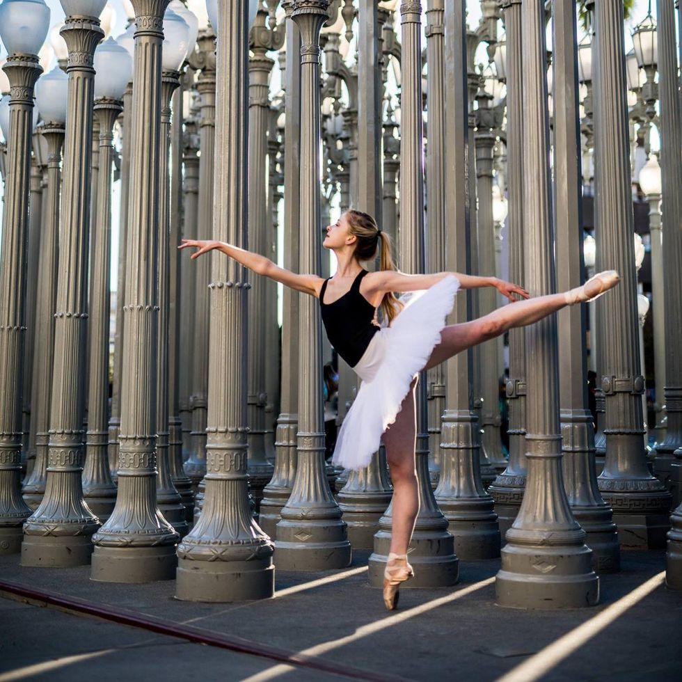 Chelasea Paige Johnston poses in first arabesque on pointe, wears a black tank leotard and white practice tutu